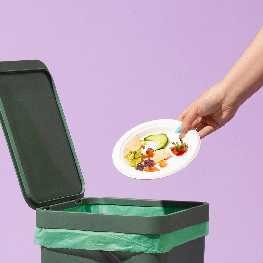 A hand shown throwing a paper plate with food on it into a green garbage bag and can